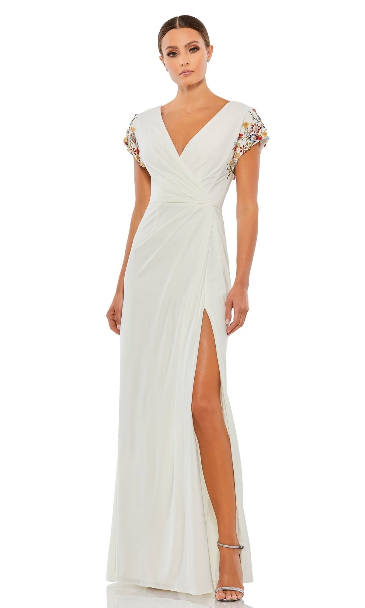 A woman stands in silver heels, wearing the Cap-Sleeve Long Formal Gown: Mac Duggal 55709, a white dress with a slit, V-neckline, and colorful embellished cap sleeves.